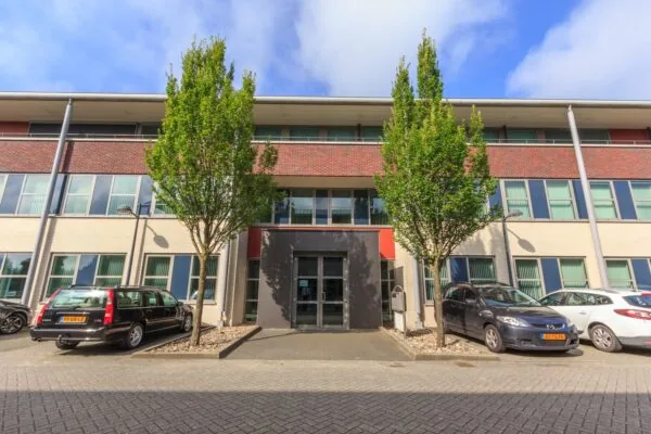 Vooraanzicht van het kantoorgebouw De Wel met bomen en geparkeerde auto's ervoor.